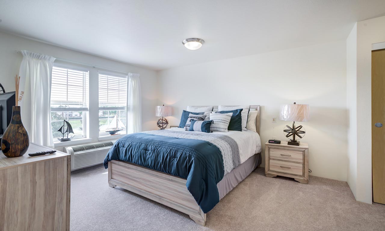 A bright, modern bedroom with a large bed adorned with blue and white bedding. Two nightstands with matching lamps flank the bed. A dresser with decorative items is positioned on the left. A window with white curtains illuminates the room with natural light.