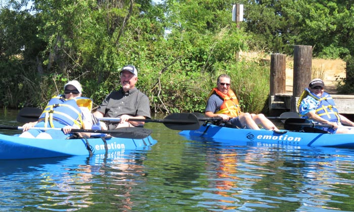 Four people are sitting in blue kayaks on a calm body of water. All are wearing life jackets, and two are wearing hats. The background includes lush greenery and wooden posts by the shore. The kayaks have 