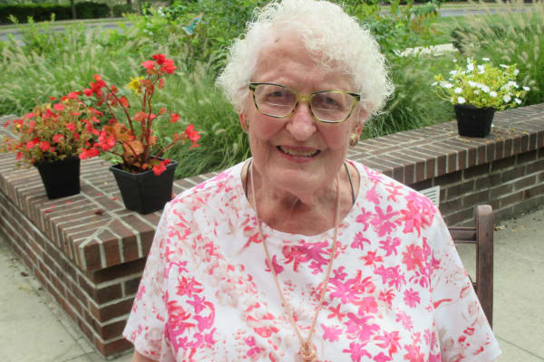 An elderly woman with curly white hair and glasses is smiling while sitting outdoors. She is wearing a white shirt with pink floral patterns. Behind her, there are brick planters with red and white flowers and green foliage.