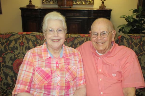 An elderly couple, both with glasses, smile while sitting closely on a patterned couch. The woman wears a pink plaid shirt, and the man wears a plain pink shirt. They are indoors, with a wooden cabinet and plant in the background.