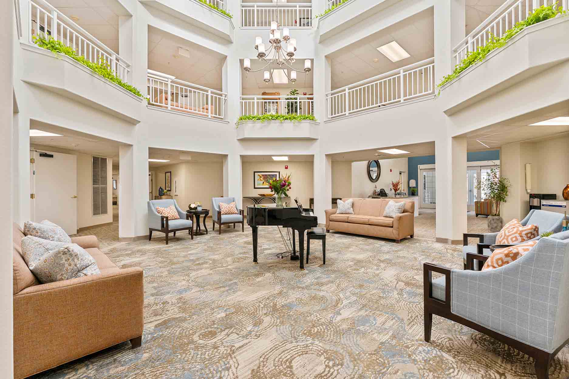 An elegant, airy atrium features a high ceiling and natural light, with green plants on the upper level railings. The space is furnished with multiple seating areas, including sofas and chairs, and a grand piano as the centerpiece atop a patterned carpet.