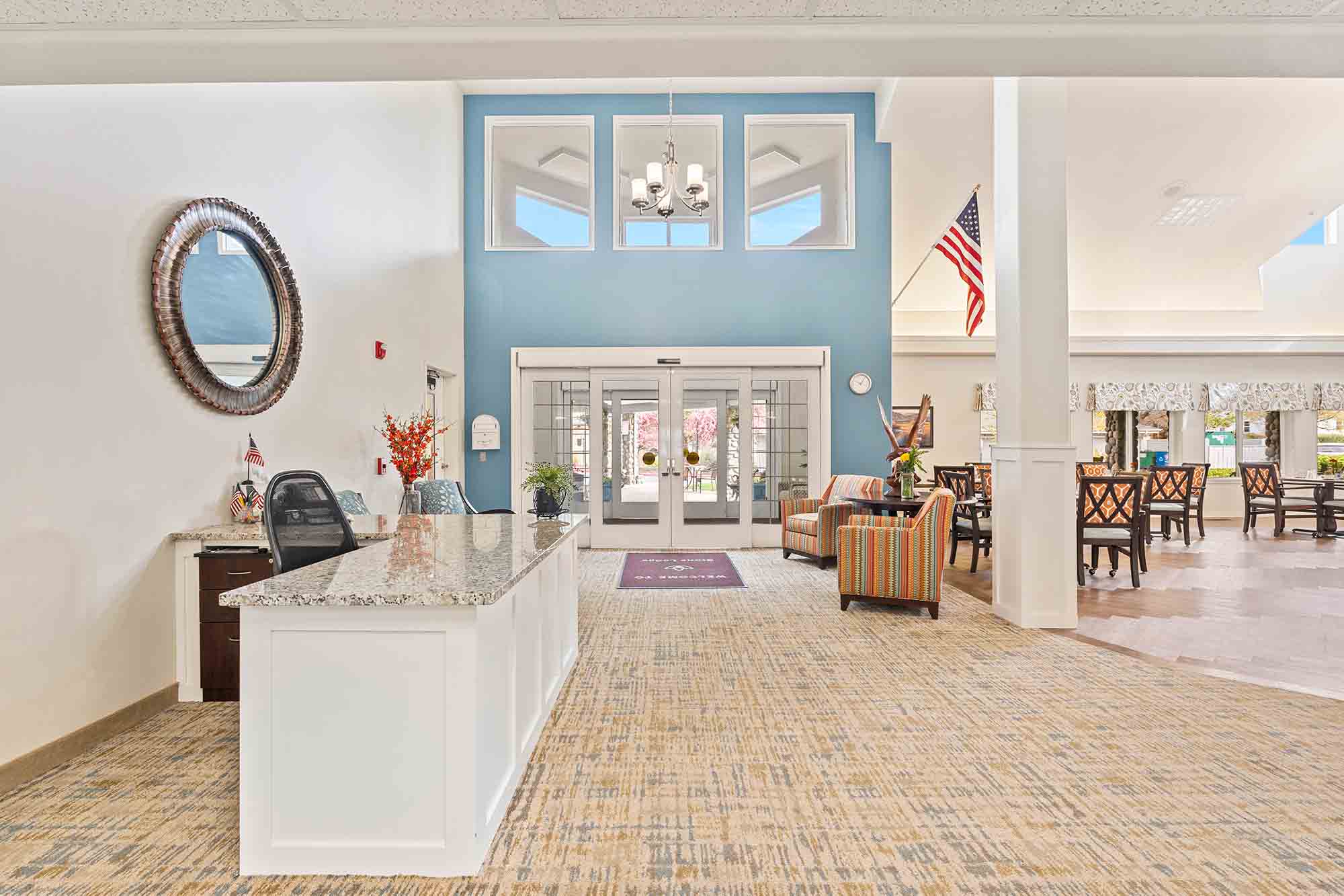 A spacious, well-lit lobby with a high ceiling. Features a front desk with a granite countertop on the left, white double doors at the entrance in the center, and seating areas with sofas and tables on the right. An American flag hangs in the background.