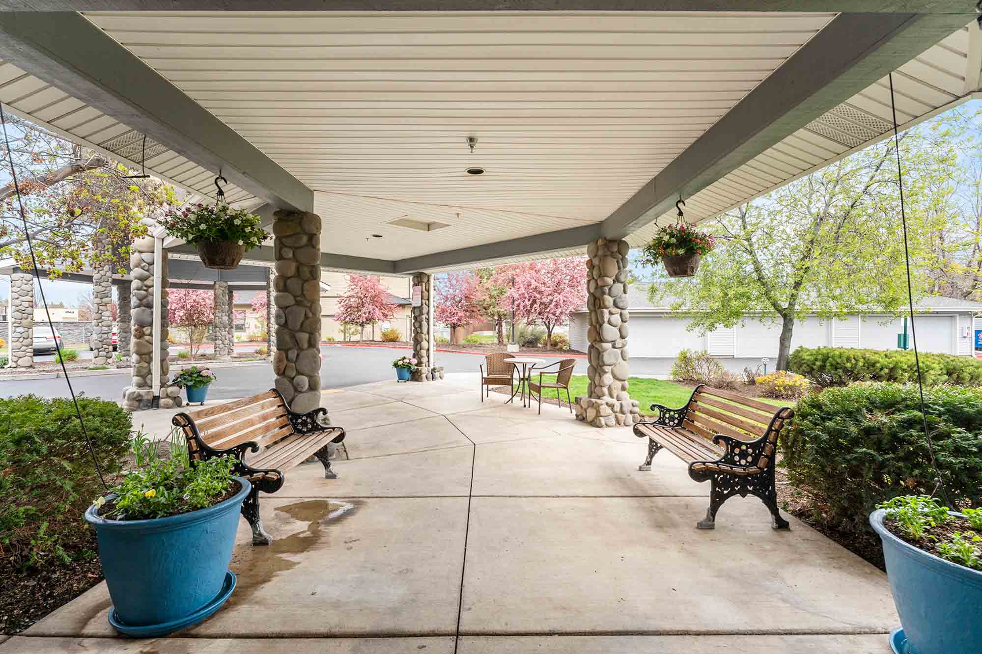 A covered outdoor area with stone pillars and a wooden ceiling, featuring two wooden benches with black metal frames. Hanging flower baskets and large blue planters with greenery decorate the space. Blooming trees are visible in the background on a sunny day.