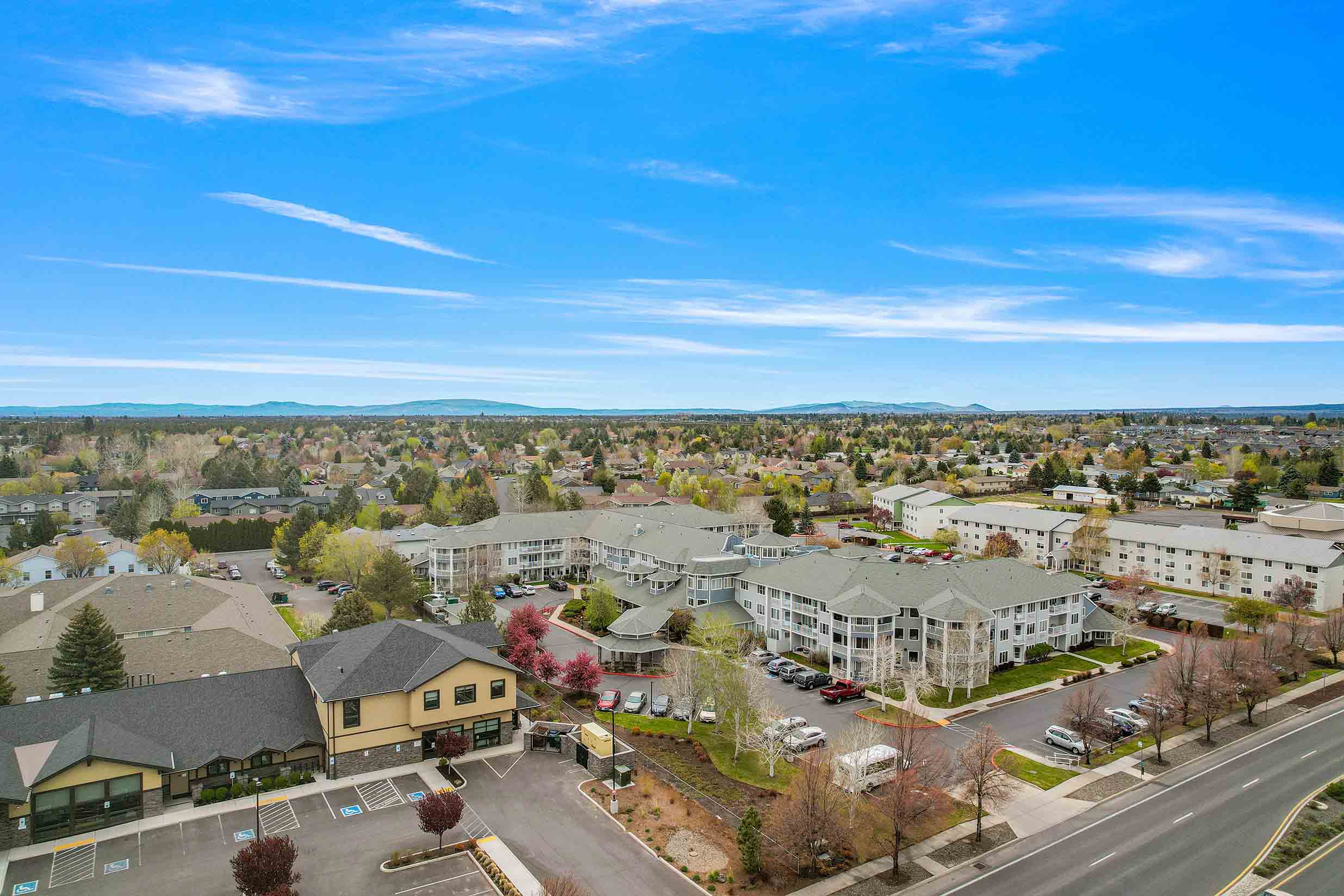 Aerial view of a suburban neighborhood with a mix of single-family homes and apartment complexes surrounded by trees. The image shows a clear day with a blue sky and scattered clouds, and the neighborhood streets are lined with parked cars and greenery.