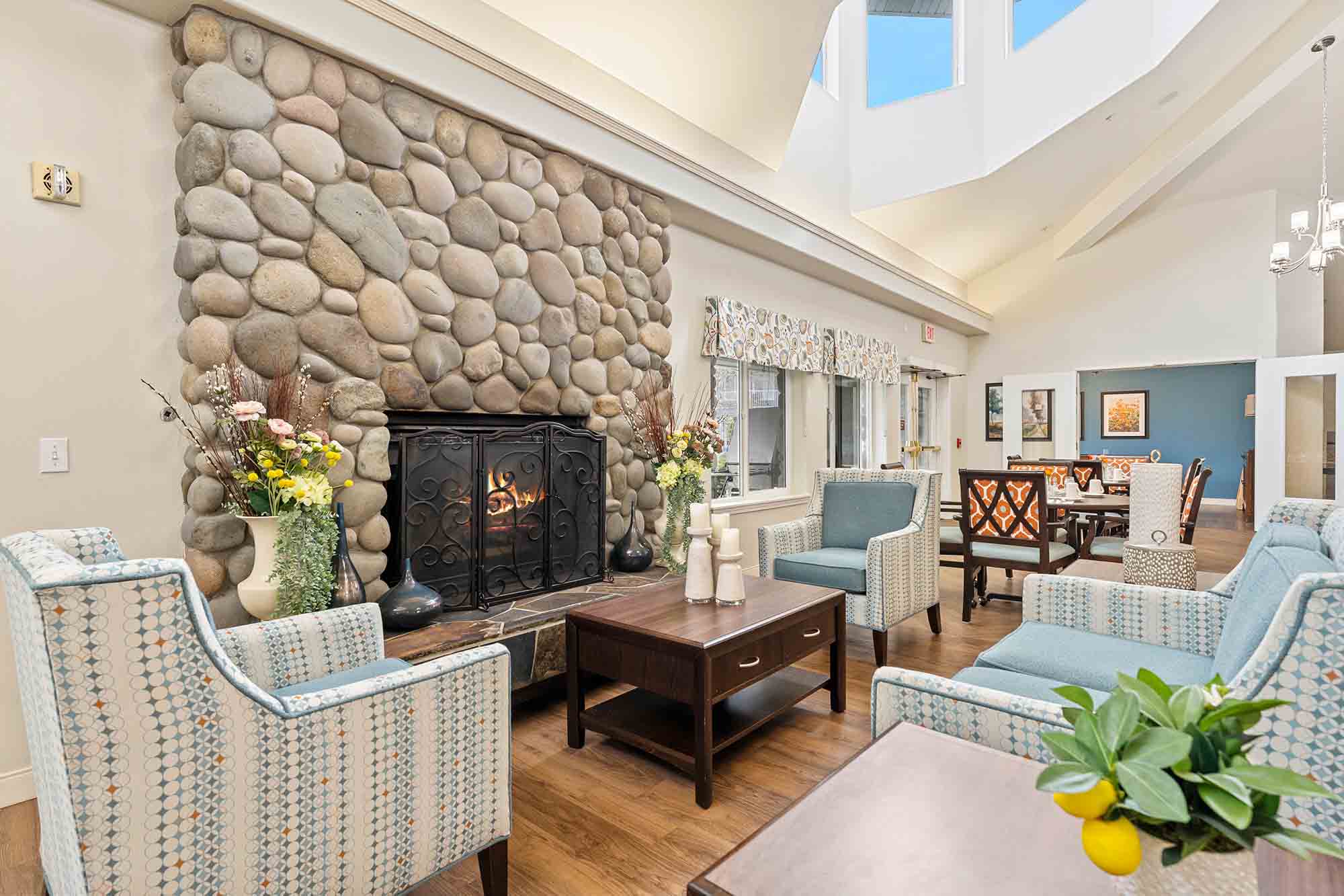 A cozy living room with a stone fireplace is surrounded by light blue armchairs and a wooden coffee table. Floral arrangements decorate the room, and large windows allow ample natural light. A dining area is visible in the background.