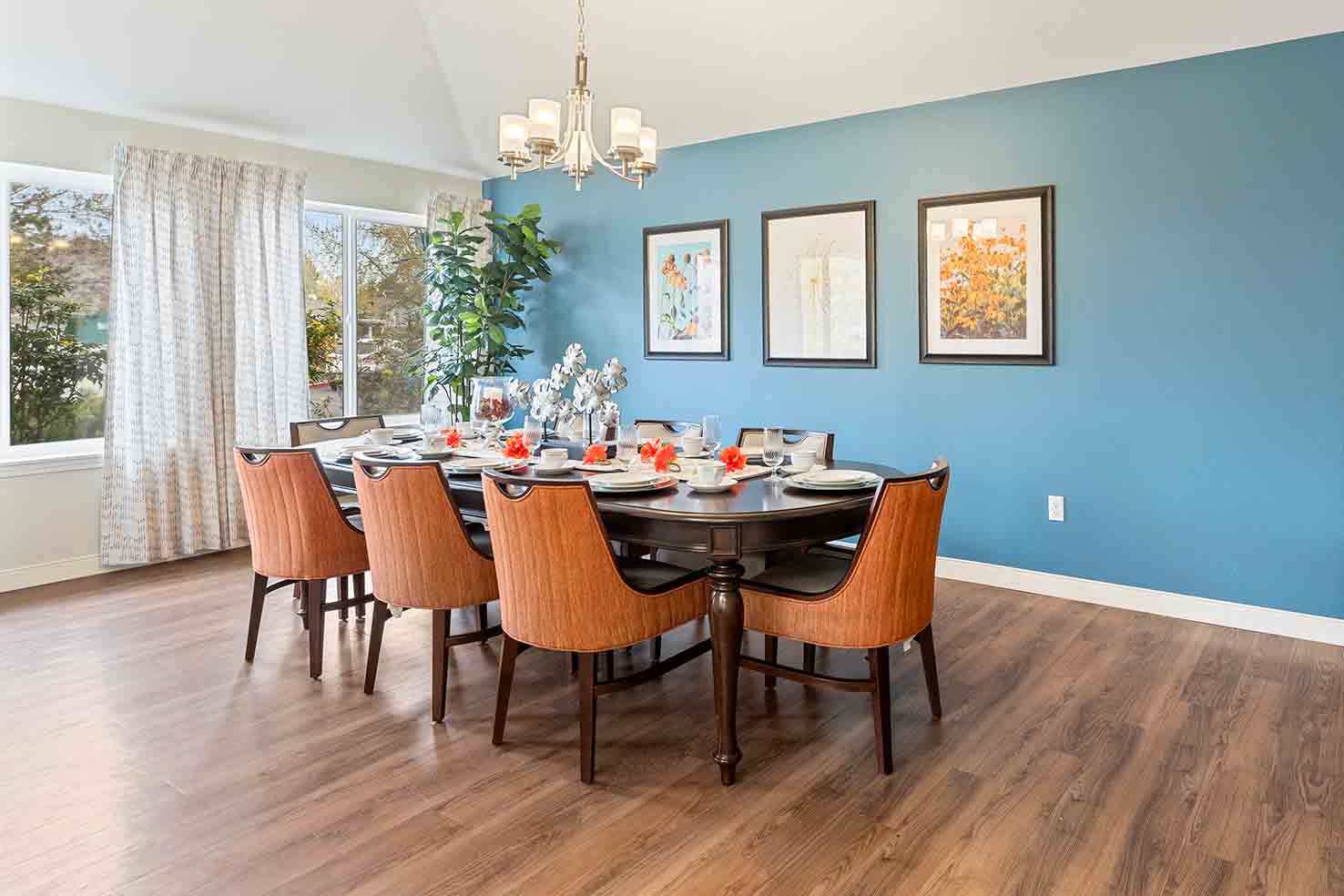 A dining room with a long oval table set for eight. The room features orange cushioned chairs, a blue accent wall with three framed paintings, hardwood floors, and a window with sheer curtains letting in natural light. A chandelier hangs above the table.