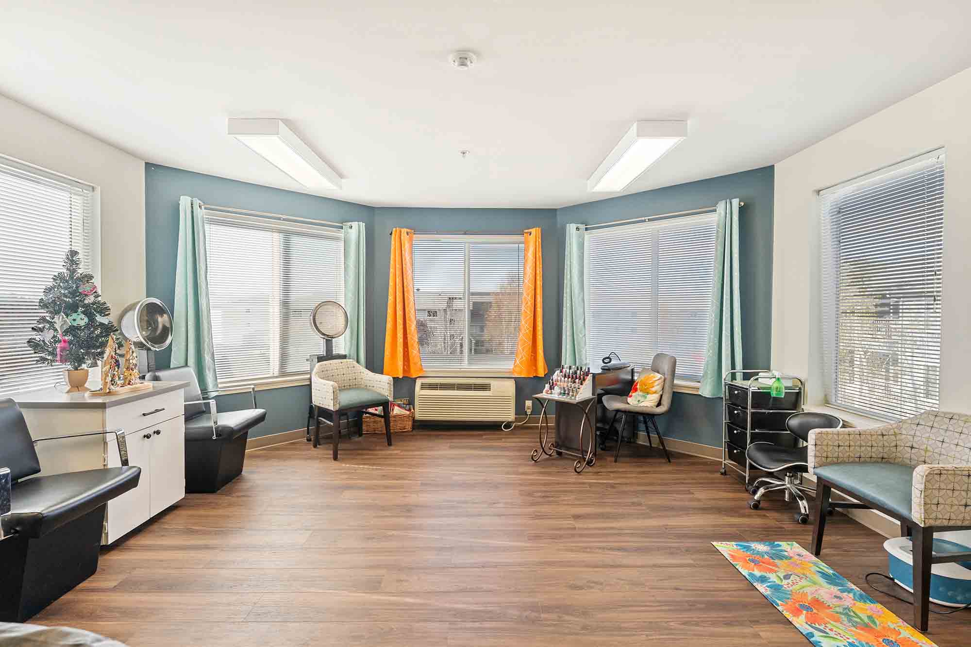 A bright, spacious room with large windows covered by light blue and orange curtains. The room has salon equipment, including chairs, mirrors, and a nail care station. The wooden floor and neutral walls create a cozy atmosphere. A small Christmas tree is seen on the left.