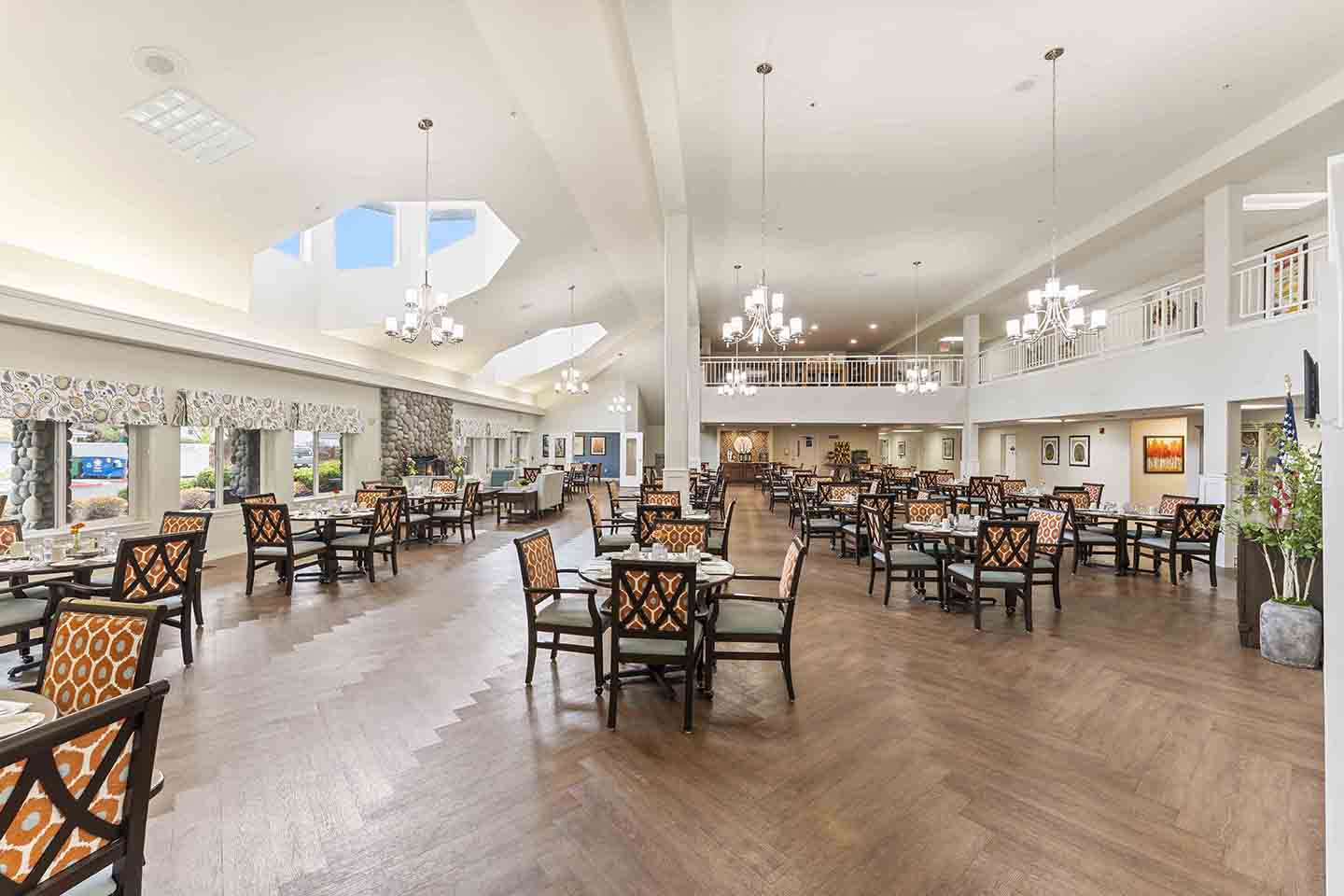 A spacious dining area with high ceilings and elegant decor, filled with numerous tables and chairs arranged neatly. Chandeliers hang from the ceiling, providing ample lighting. Large windows on the left allow natural light to brighten the room.