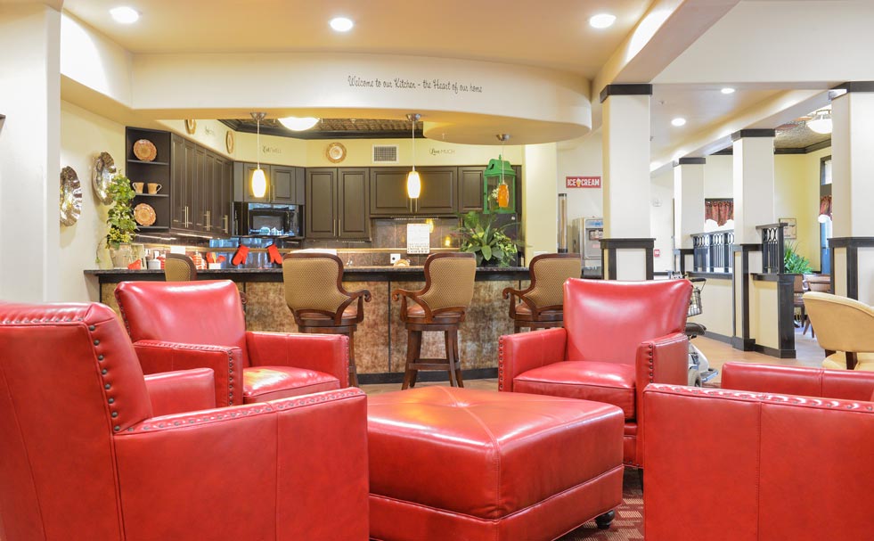 A cozy and inviting room features four red leather armchairs and a matching ottoman arranged in a square around a coffee table. The background reveals a kitchen with dark cabinets, barstool seating, and warm ambient lighting. A sign above the kitchen reads 