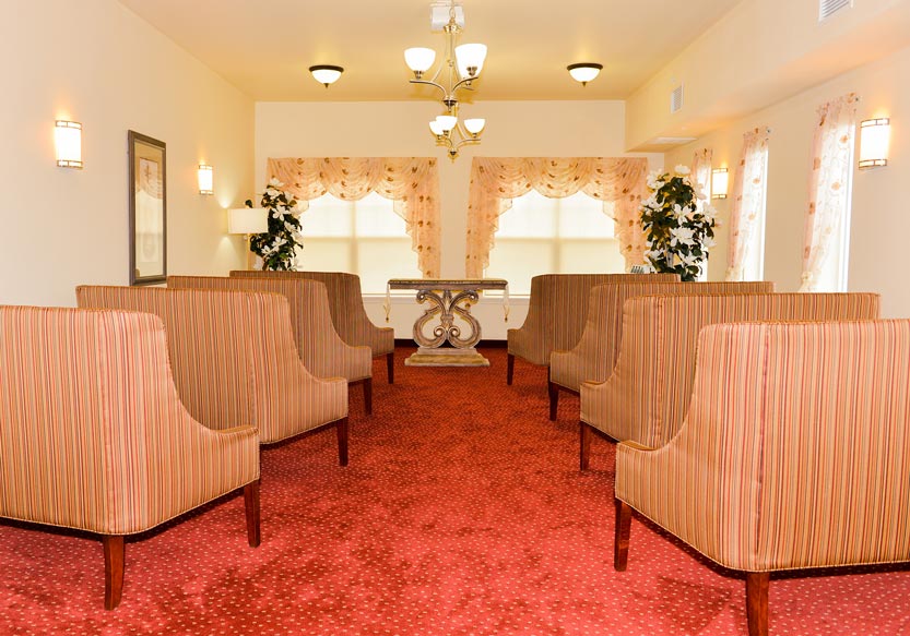 A small, well-lit chapel with red carpeting and beige walls, featuring ten striped upholstered chairs arranged in two rows. The room has large windows with decorative drapes, wall sconces, potted plants, and a small ornate table at the front.