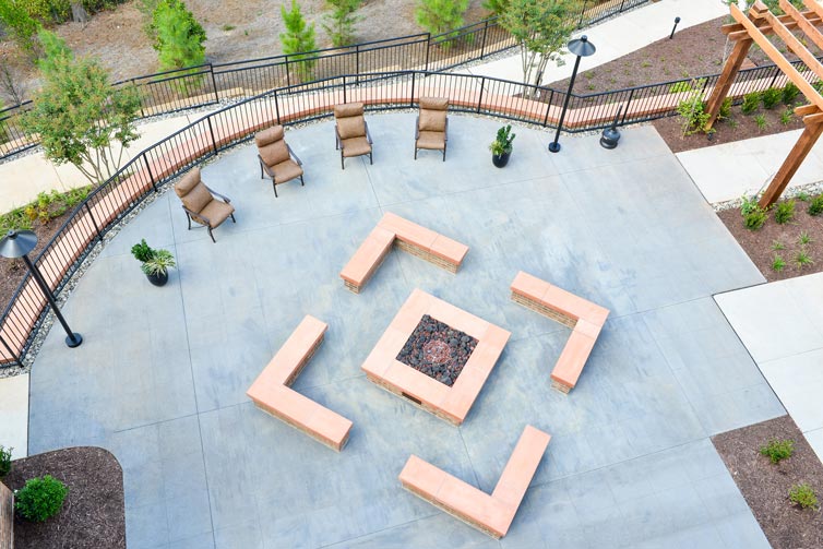 Aerial view of an outdoor seating area featuring a square fire pit surrounded by cushioned benches arranged in a square pattern. Several recliner chairs are aligned along a curved pathway, with lampposts, potted plants, and greenery visible in the background.