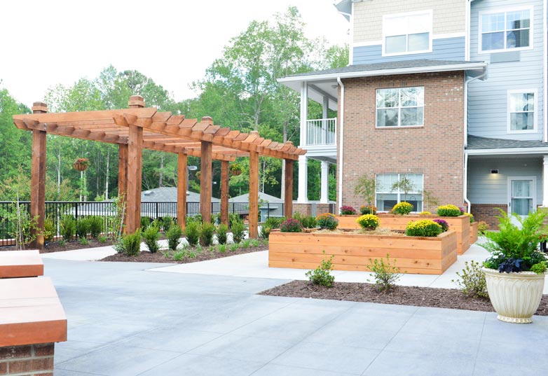 An outdoor patio area with a wooden pergola, surrounded by raised garden beds filled with colorful flowers. In the background, there is a multi-story residential building with a red brick and siding exterior. Trees are visible in the distance.