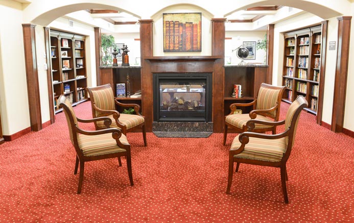 A cozy library scene with a red carpet featuring four striped upholstered chairs arranged around a modern fireplace. Bookshelves line the walls, filled with books. A framed painting of books hangs above the fireplace.