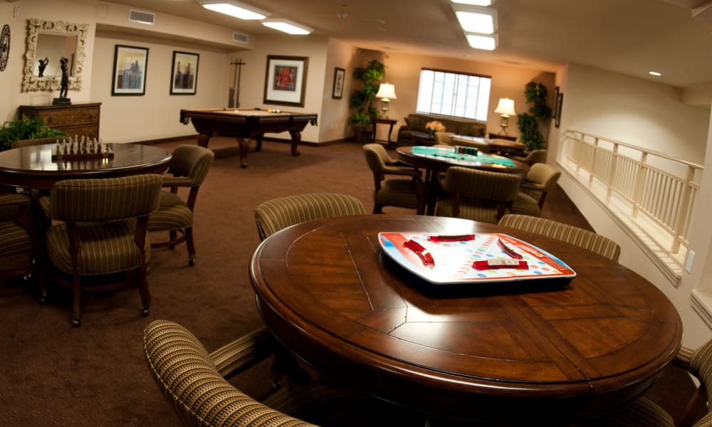 A well-lit recreation room featuring several round tables with board games, including Scrabble. There are also pool tables, framed wall art, and comfortable seating with striped chairs. The flooring is carpeted, and natural light streams in from a window in the back.