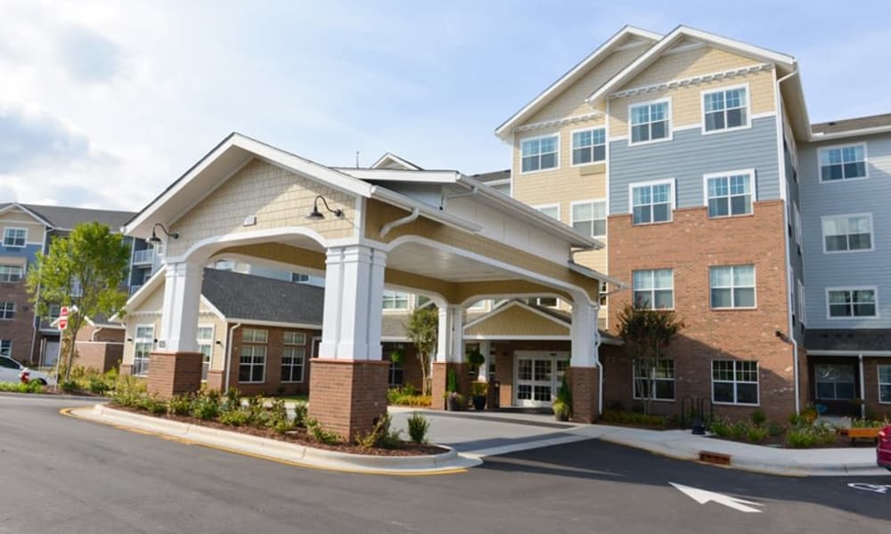 A modern multi-story building with a brick and panel exterior features a covered entrance with white pillars. The surrounding area is well-maintained with landscaping, and the sky is partly cloudy. A driveway leads to the entrance with parking available.