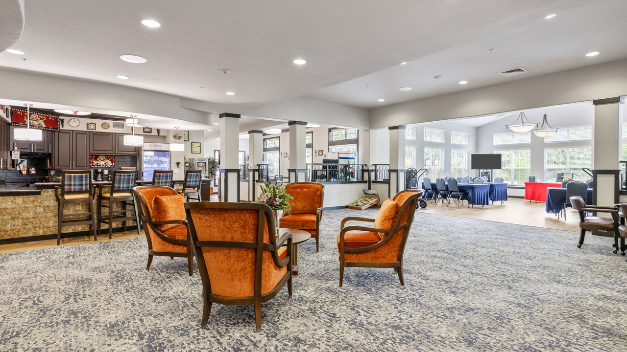 Spacious room with orange armchairs around a small table, a bar area on the left, and tables with blue and red tablecloths near large windows in the background.