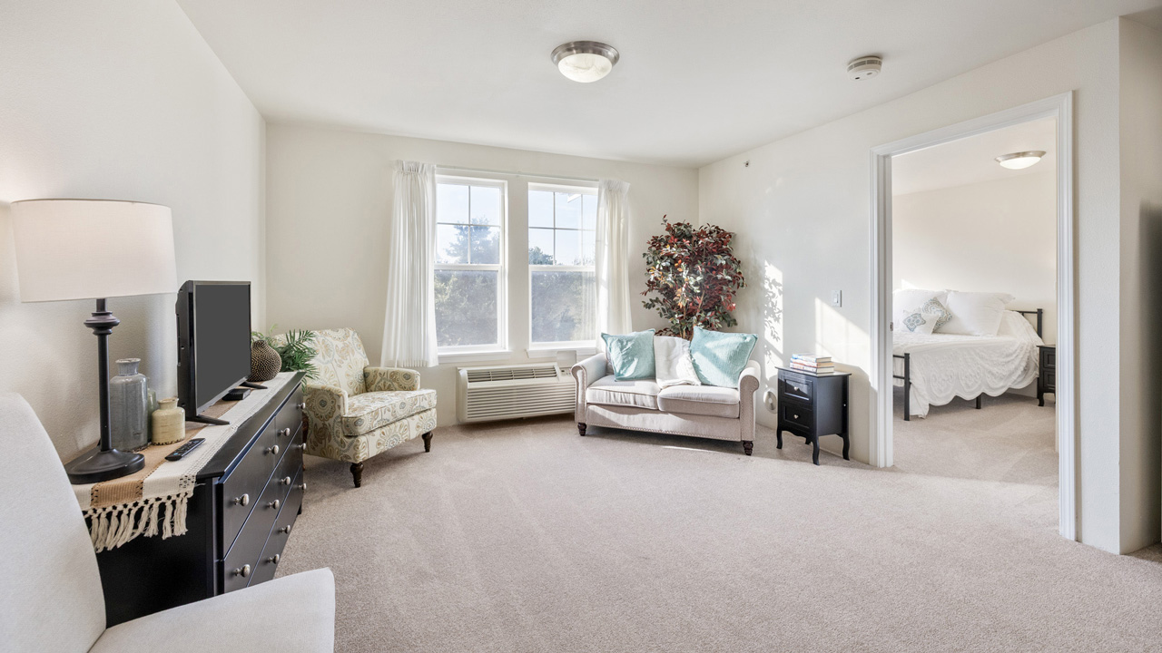 Bright living room with beige carpet, a sofa, armchairs, a TV on a dresser, a lamp, and a large window. An open doorway leads to a bedroom with a made bed.