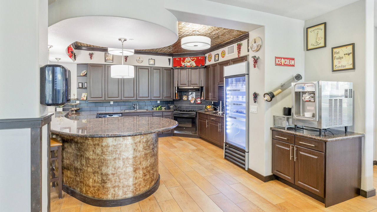 Modern kitchen with dark cabinets, granite countertops, a large curved island, wood floors, a beverage machine, wall decor, and overhead lighting. Refrigerator and microwave are visible near the back wall.