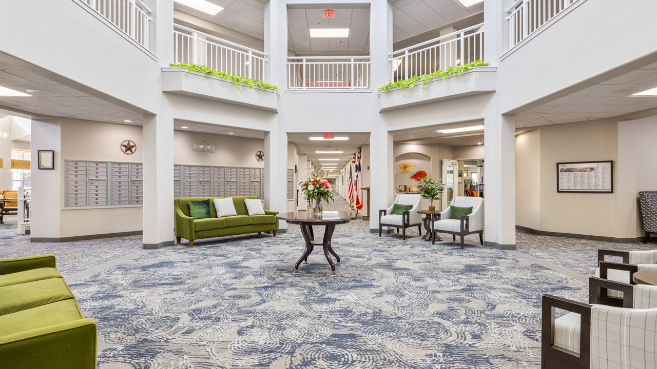 Bright lobby with patterned carpet, green and beige seating, a round table with flowers, mailboxes on the left, flags down a hallway, and second-floor balconies with plants. Neutral walls and ample natural light.