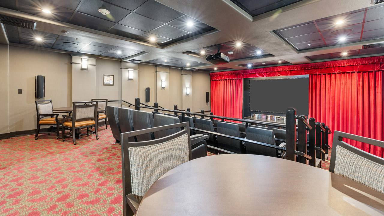 A small movie theater with tiered seating, a round table with chairs in the foreground, red carpet, and a large screen framed by red curtains. Ceiling and walls have modern lighting and speakers.