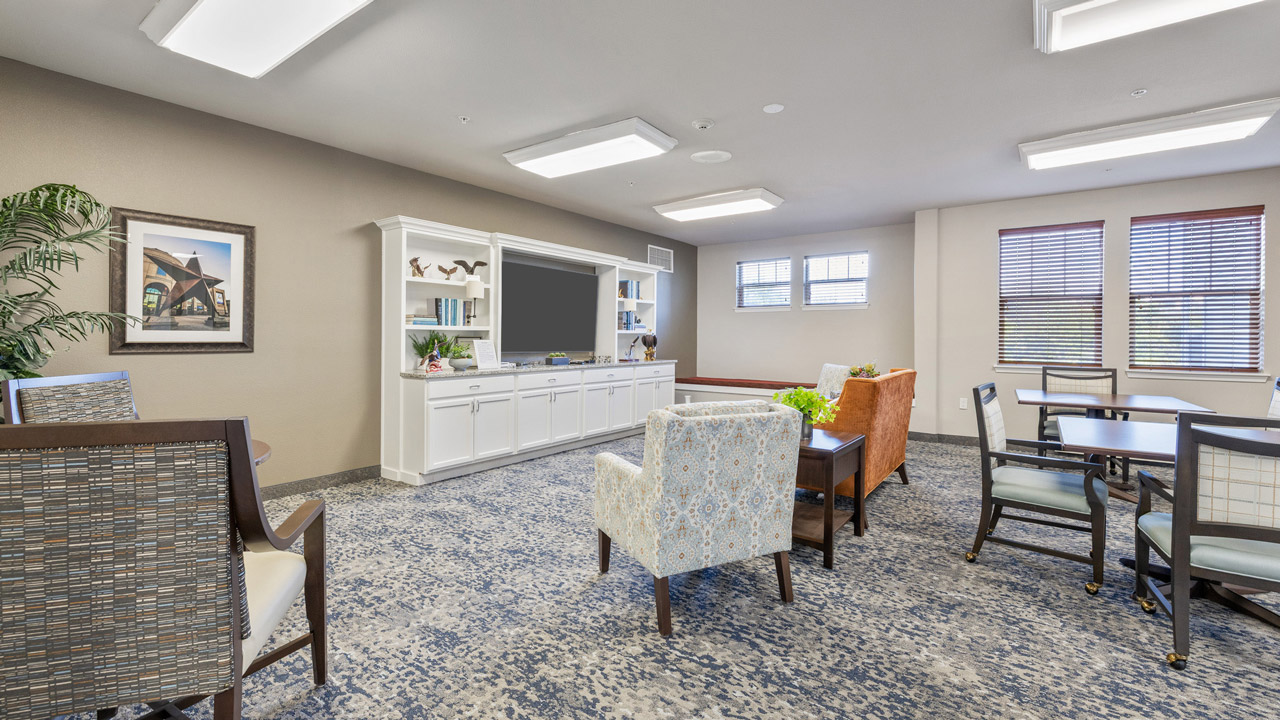 A bright, modern lounge with patterned carpet, various upholstered chairs, a white entertainment center, framed artwork, potted plant, and windows letting in natural light.