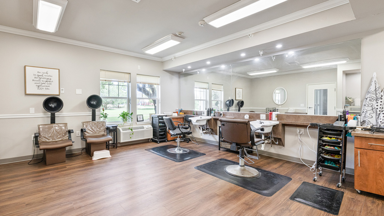 A bright, tidy salon with two styling chairs, two hair drying stations, a large wall mirror, and organized shelves, all on wood flooring with natural light coming through the windows.