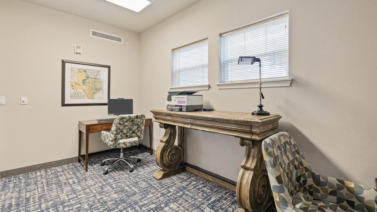 Small office with a patterned carpet, two chairs, a wooden desk with a printer and lamp, a second desk with a computer, Texas map on the wall, and two windows letting in natural light.