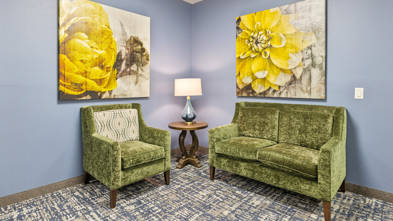 A small room with a green armchair and loveseat, a round wooden table with a lamp, and two large yellow floral paintings on blue walls. Blue-patterned carpet covers the floor.