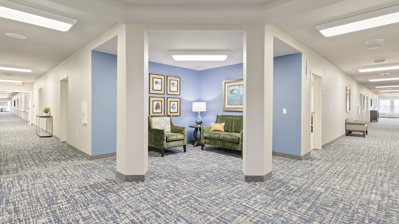 Bright hallway with patterned blue carpet, off-white walls, and a cozy nook featuring two green chairs, a lamp, and framed artwork. Several doors line the corridor, and ceiling lights illuminate the space.