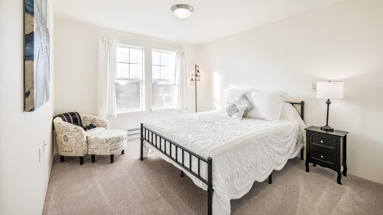Bright bedroom with a metal-framed bed, white bedding, two accent pillows, a black nightstand with a lamp, an armchair with script fabric, large windows with white curtains, and beige carpet flooring.