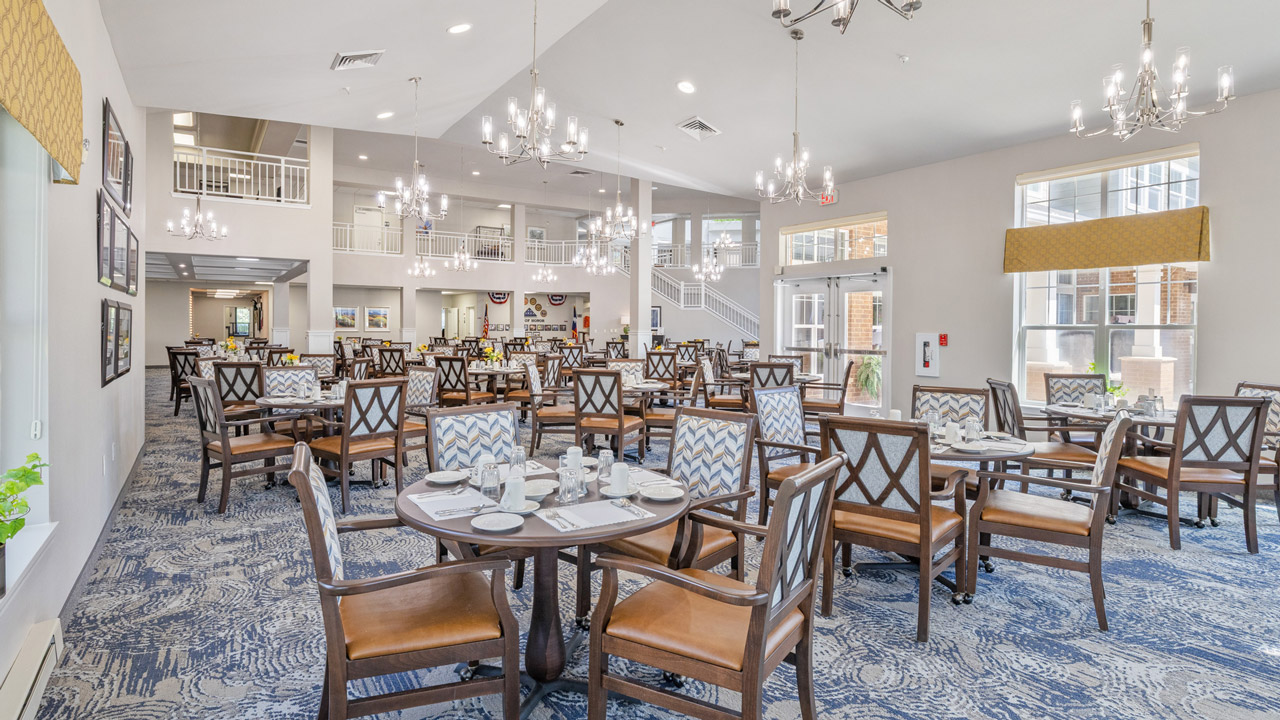 A spacious, well-lit dining room with neatly set tables and wooden chairs, chandeliers, large windows, and patterned carpet. The room has a clean, elegant atmosphere with neutral tones and modern decor.