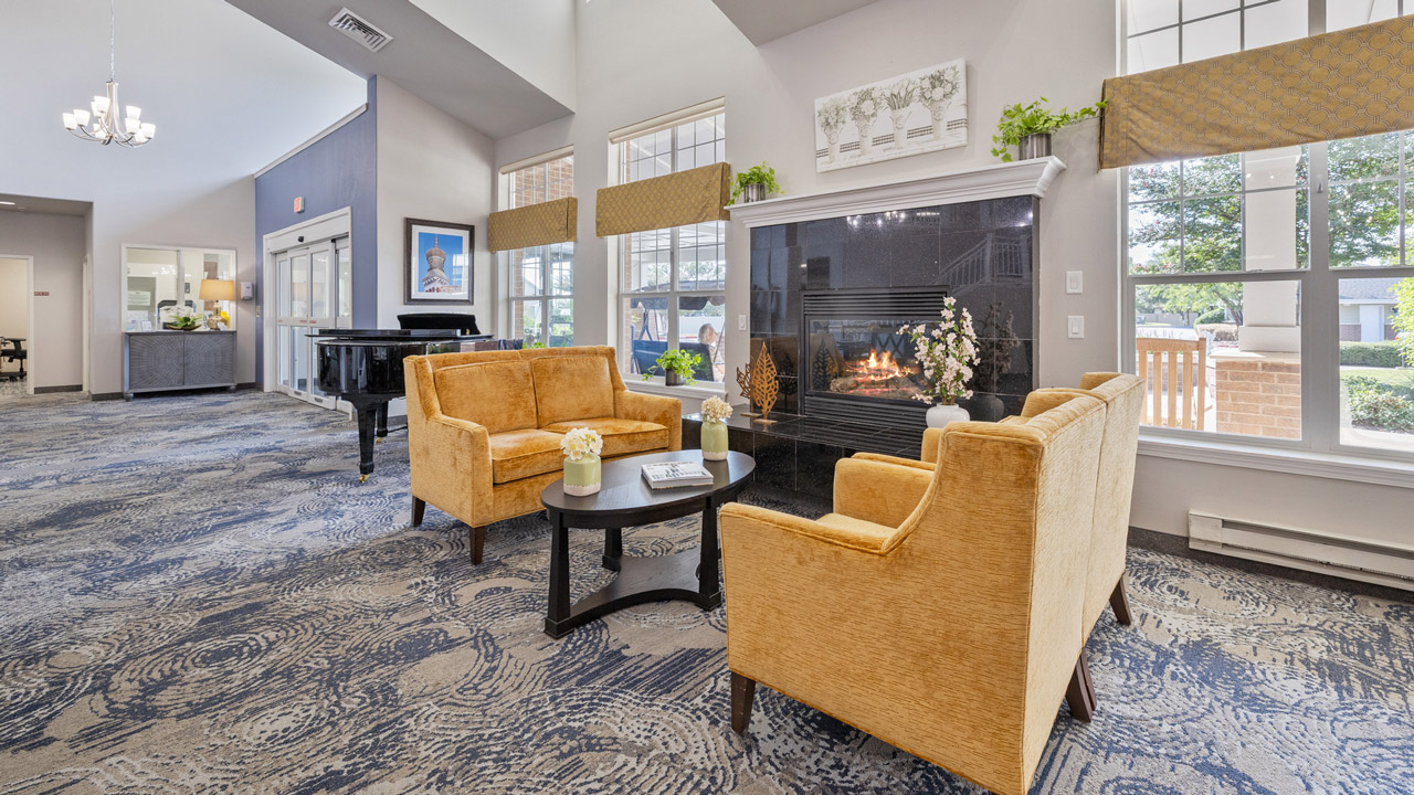 A bright, modern lounge with yellow armchairs, a black piano, and a lit fireplace. Large windows let in natural light. There are decorative plants and a coffee table with books and flowers.