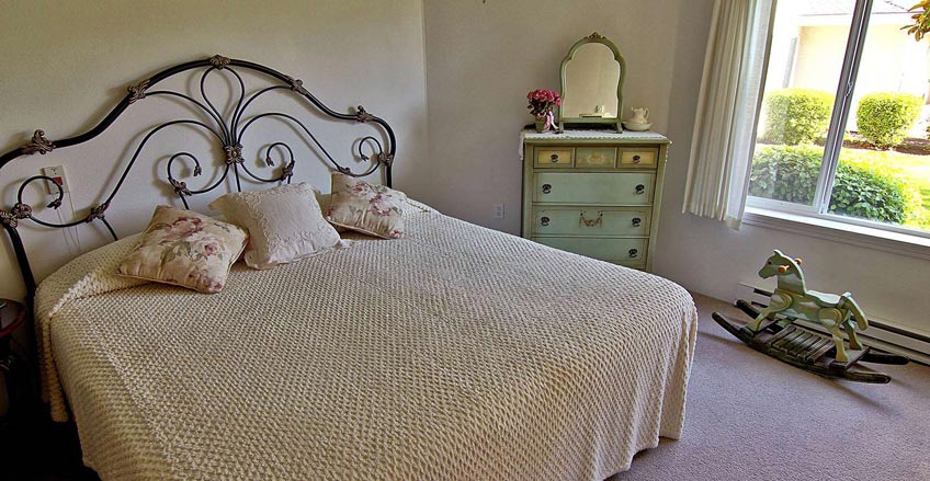 A cozy bedroom features a large bed with an ornate metal headboard and floral pillows. A vintage dresser with a mirror sits against the wall, adorned with decorative items. A rocking horse rests by the window, which offers a view of a well-maintained garden.