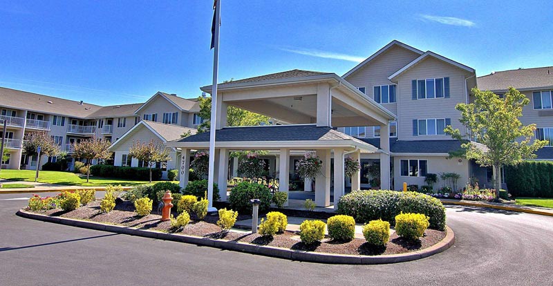 Two-story residential building with beige walls and white-trimmed windows, surrounded by well-maintained landscaping with shrubs and flowers. The entrance features a large, covered porch. The sky is clear and blue, and a fire hydrant is visible near the roundabout driveway.