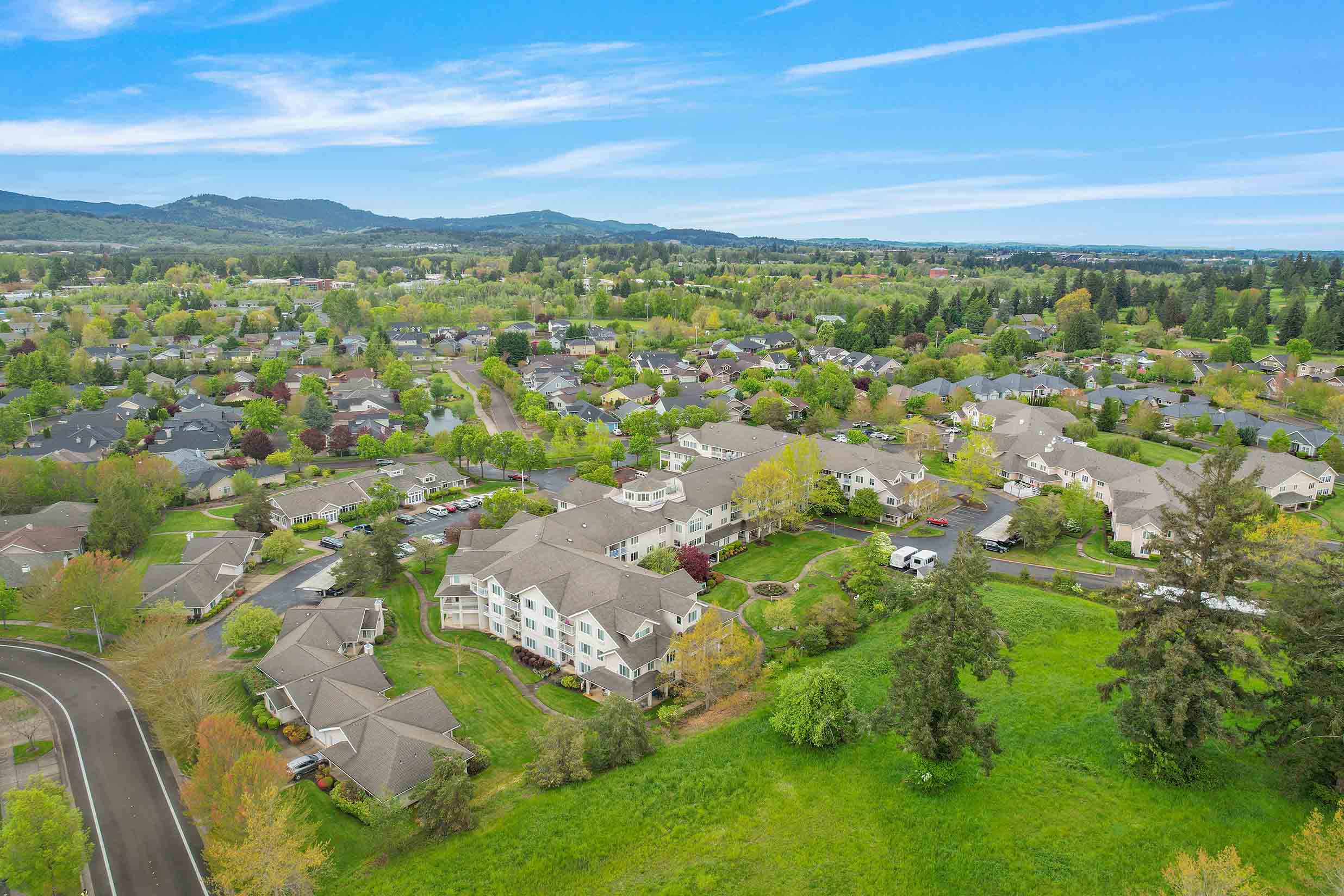 Aerial view of a suburban neighborhood featuring numerous houses surrounded by lush green trees and open grassy areas. In the background, rolling hills are visible under a partly cloudy blue sky. The area appears peaceful and well-maintained.