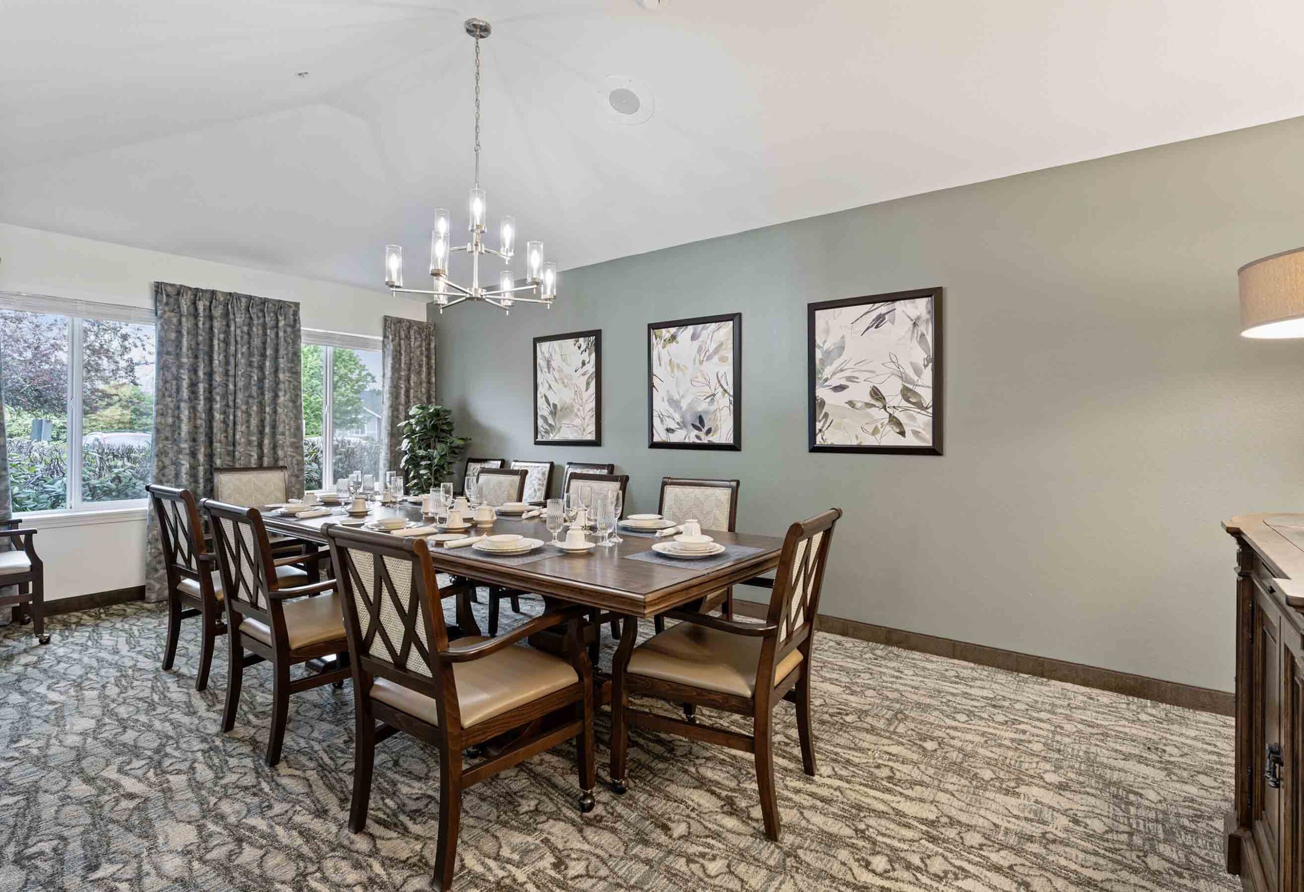 A dining room with a long wooden table set for eight, featuring cushioned chairs, a chandelier above, and decorated walls with three framed botanical prints. Large windows with curtains allow natural light to enter, and a sideboard with a lamp stands to the right.