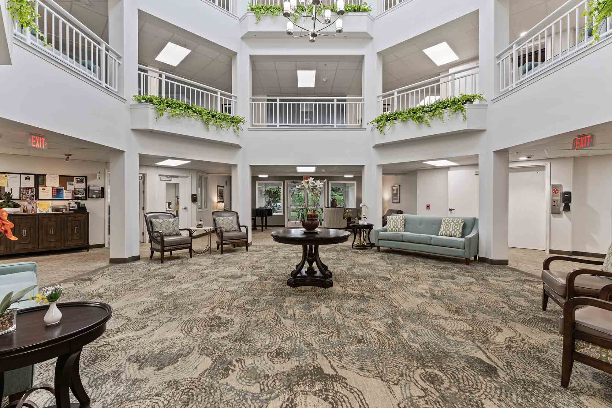 A spacious, well-lit atrium with three levels. The ground floor features a large carpeted area with a round table in the center and furniture including sofas, chairs, and side tables. Green plants adorn railings on the upper levels. Double doors are visible at the back.