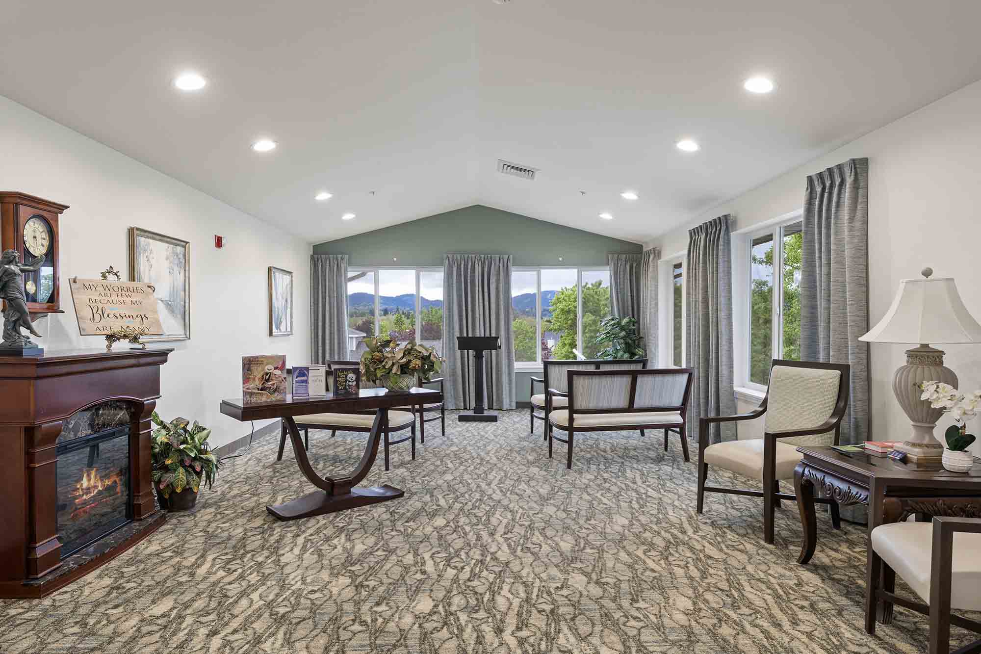 A cozy living room features a fireplace with a clock above it, a small wooden table with decorations, comfortable chairs, and large windows with views of trees and mountains. The room has patterned carpet, light gray curtains, and potted plants.