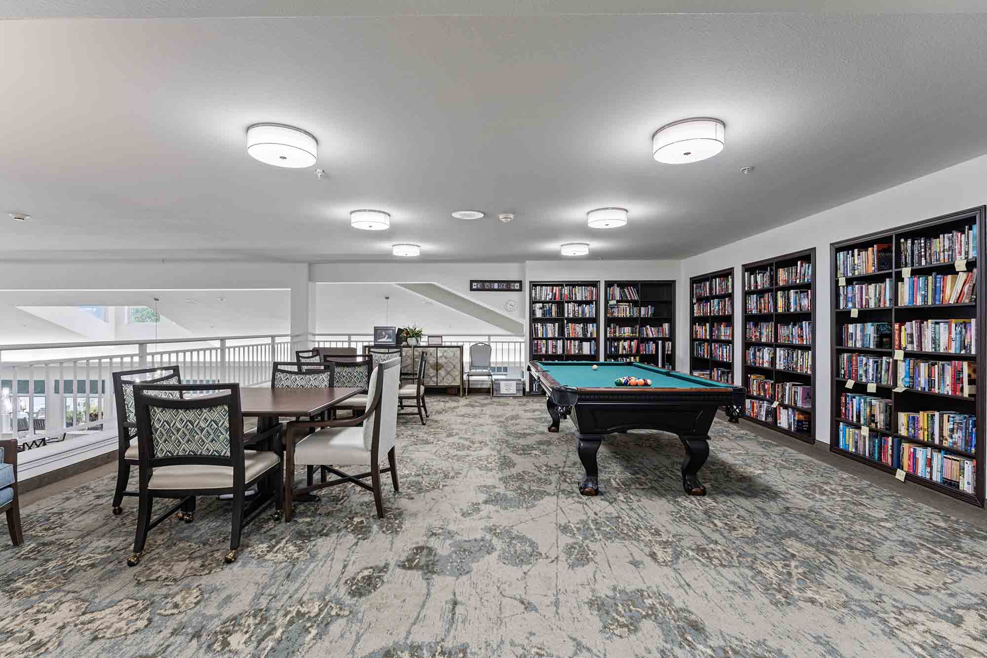 A well-lit library room featuring a billiards table in the center, multiple bookshelves filled with books along the walls, and a table with chairs for reading or games. The room has a cozy carpeted floor and a modern, spacious design.