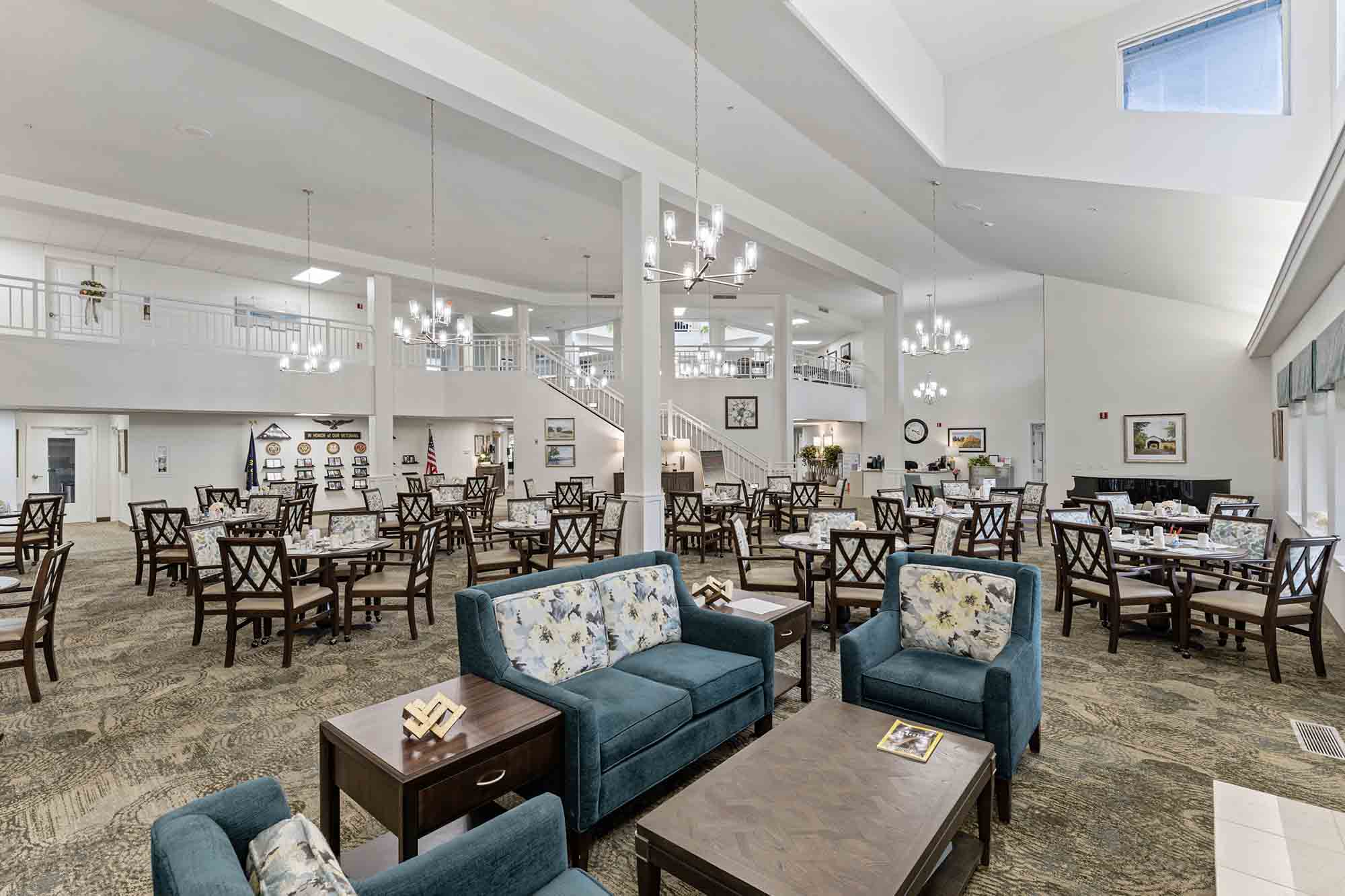 A spacious, well-lit dining area with high ceilings and modern chandeliers. The room features numerous tables set with chairs, some arranged in a more relaxed, lounge-like setup with sofas and coffee tables. A staircase and balcony are visible in the background.