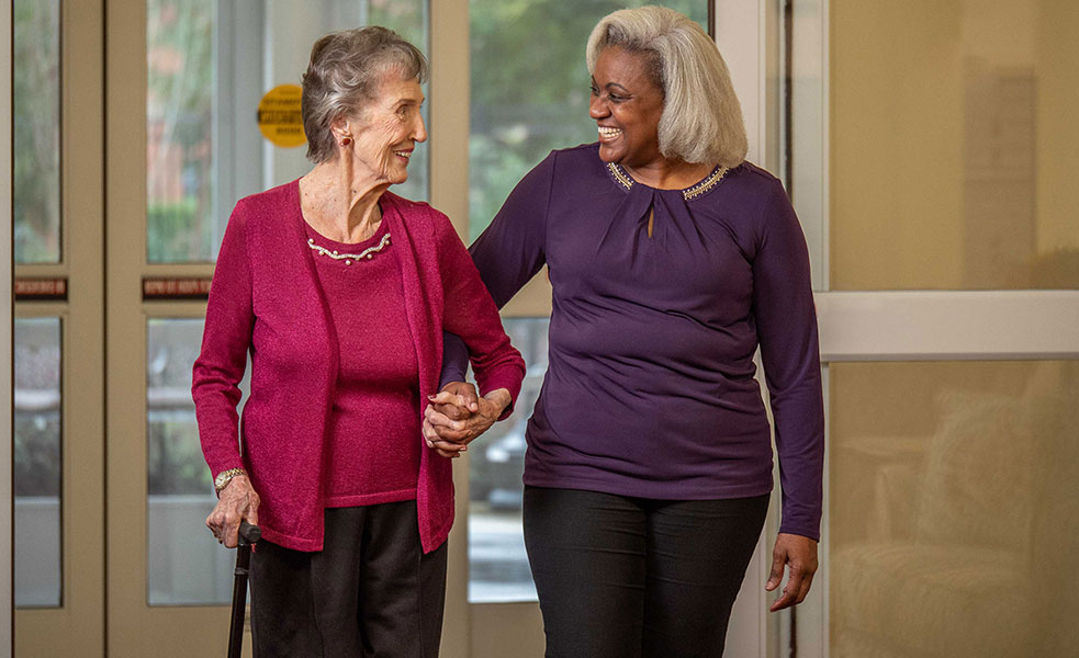 An elderly woman with a cane, wearing a red cardigan, holds hands and walks with a smiling woman in a purple top. They appear happy and engaged as they walk together indoors.