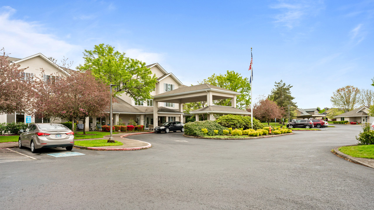 A residential or assisted living facility with a covered entrance, surrounded by trees, bushes, a flagpole, and parked cars on a paved driveway under a blue sky.