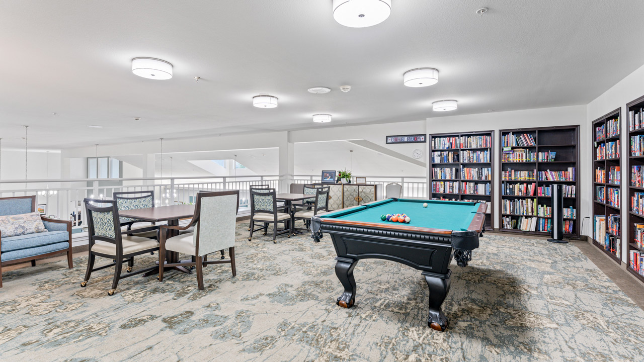 A spacious game room features a pool table with balls arranged, a card table with chairs, bookshelves filled with books, and a cozy seating area, all under bright ceiling lights on a patterned carpet.