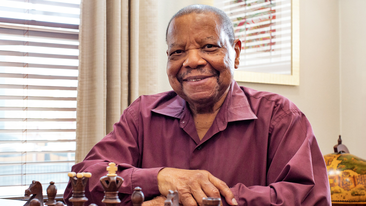 An older man with short hair and a warm smile sits at a table with a chessboard, wearing a maroon shirt, in a sunlit room with blinds and beige curtains in the background.