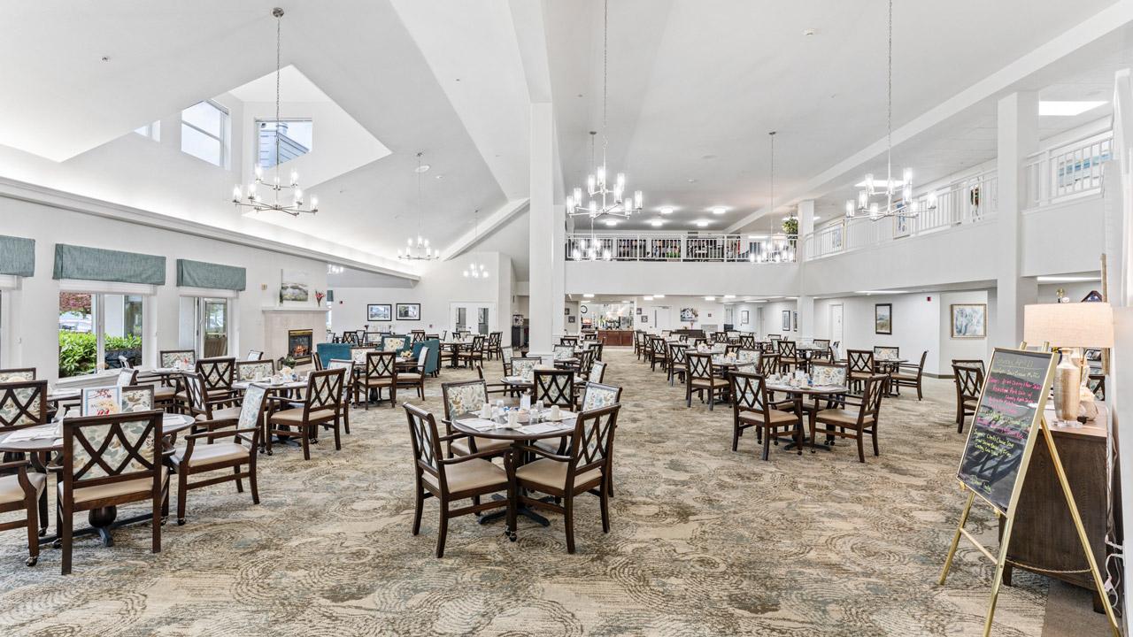 A spacious, bright dining hall with high ceilings, multiple chandeliers, and many neatly arranged tables with wooden chairs. Large windows let in natural light, and a signboard stands near the entrance.