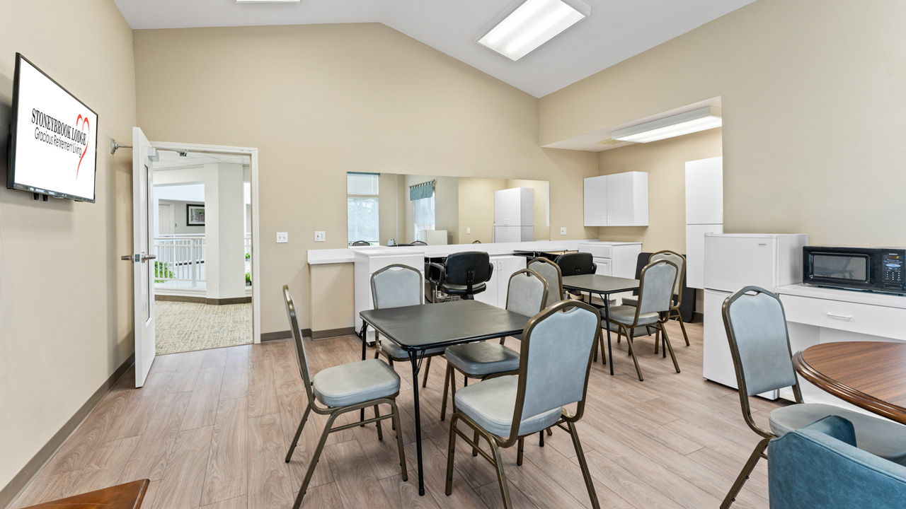 A break room with several tables and chairs, a kitchenette area with white cabinets, a microwave, and a refrigerator. A wall-mounted TV displays a heart logo and "Snohomish County" text. Walls are beige, and the floor is light wood.