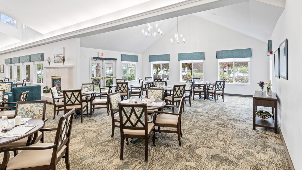 A spacious, bright dining area with several round tables and wooden chairs, large windows with teal valances, white walls, a chandelier, and decorative plants, creating a welcoming, elegant atmosphere.