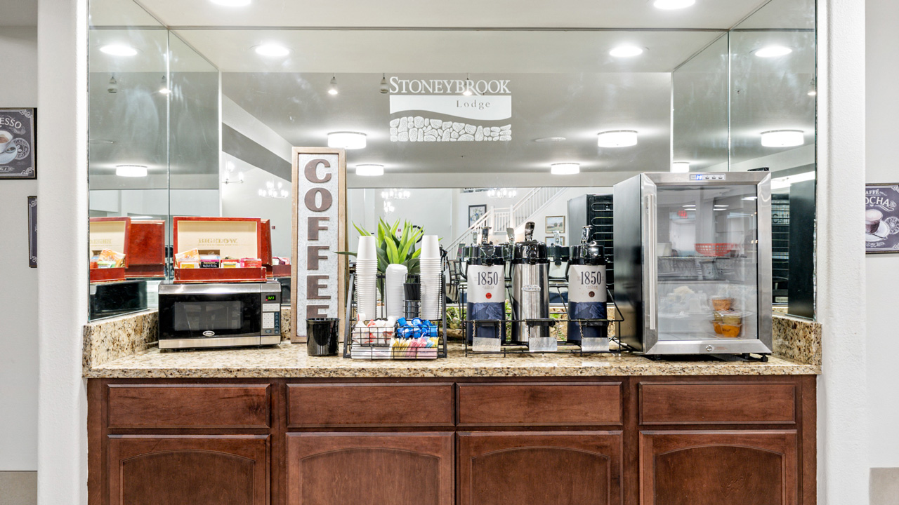 A hotel coffee station with a microwave, coffee dispensers, paper cups, creamer, sugar, napkins, and a small refrigerator on a granite countertop in front of a mirrored wall.