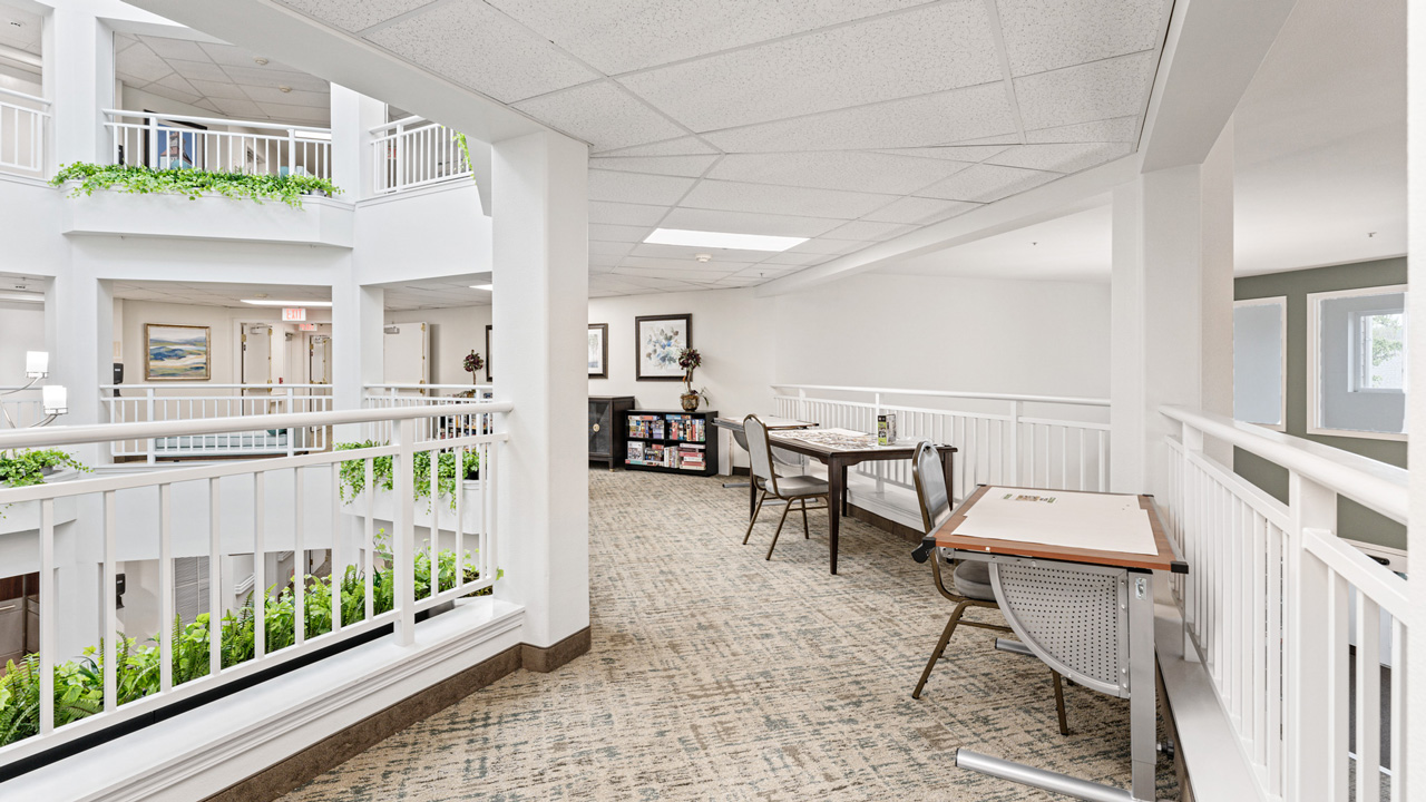 Bright, open indoor balcony area with white railings, green plants, tables and chairs for seating, carpeted floor, and framed artwork on the walls in a modern building.