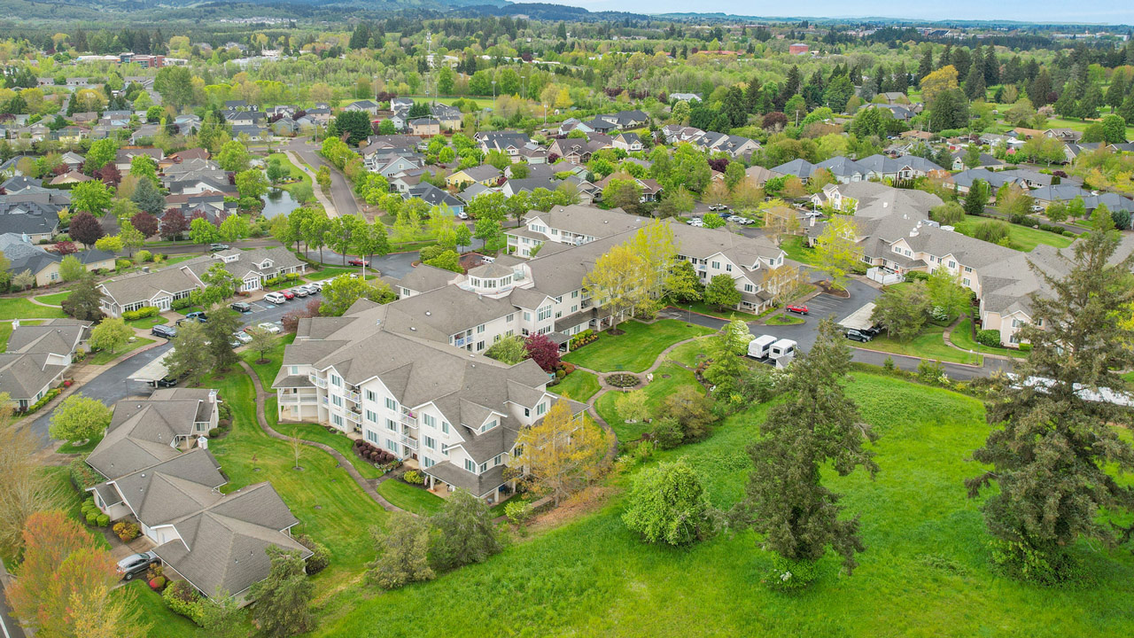 Aerial view of a well-maintained residential community with multiple apartment buildings, green lawns, trees, winding roads, and surrounding natural landscape. The area appears peaceful and suburban.